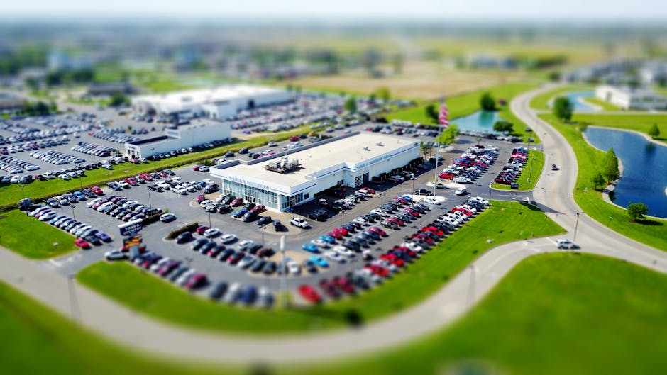 Aerial high-angle view of a bustling car dealership surrounded by parked cars in a green landscape